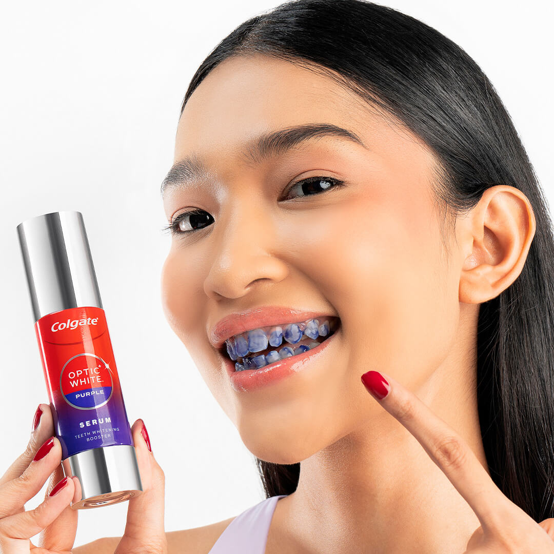 Seorang gadis muda menunjukkan botol Serum Optic White Purple dan giginya, yang telah disapu dengan serum ungu